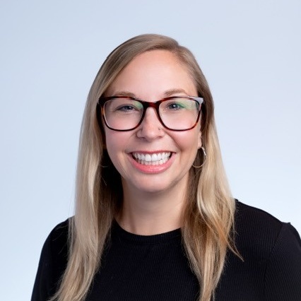 Portrait of a smiling woman with blonde hair, red-framed glasses, and hoop earrings against a light blue background.